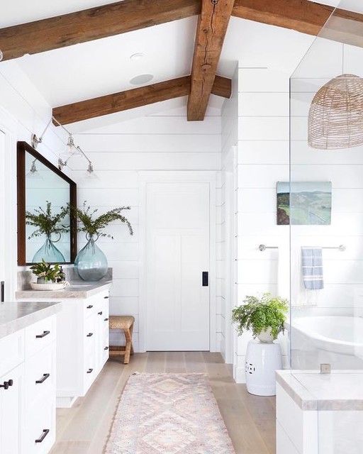 a white coastal bathroom with rich stained wooden beams, two vanities, a bathtub and a shower space plus greenery