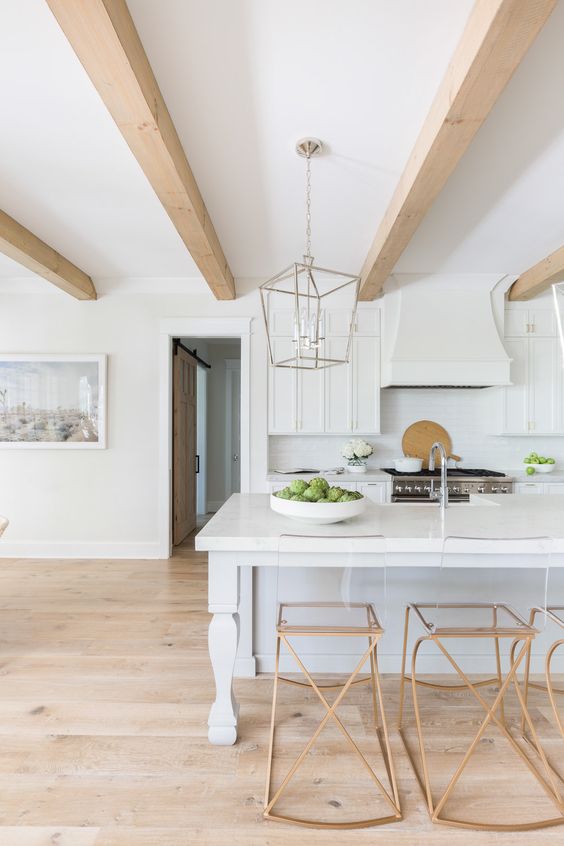 an airy white kitchen with an elegant kitchen island, copper stools, light-colored wooden beams that add warmth to the space