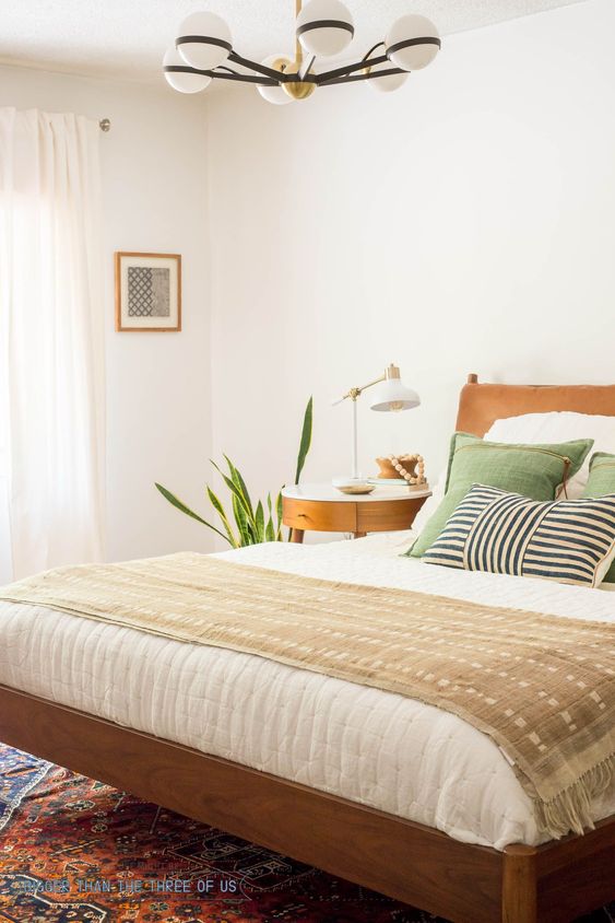 a cozy mid-century modern bedroom with white walls, warm-colored furniture, potted plants, a chandelier with globes and green pillows