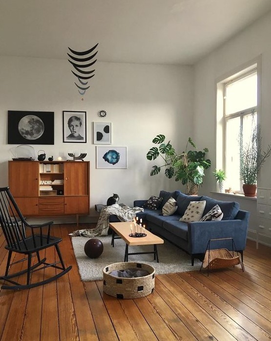 a mid-century modern living room with a blue sofa, a stained buffet, a black rocker, a low bench, a basket for storage and a cool gallery wall