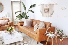 a mid-century modern to boho living room with tan leather furniture, a low white coffee table, woven poufs, a macrame hanging and potted plants