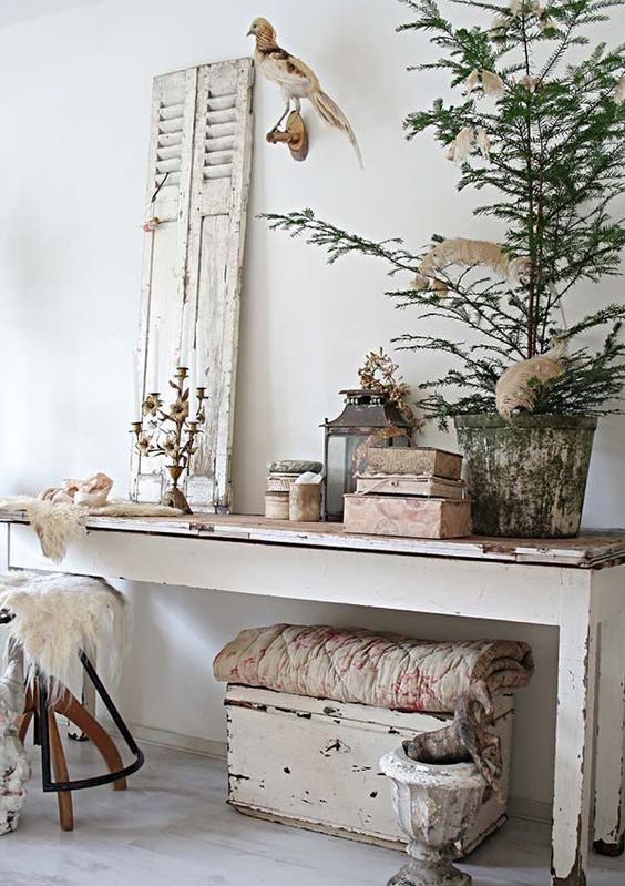 a shabby chic entryway with a long console table, a chest under it, a stool, a shutter, candelabras and candle lanterns