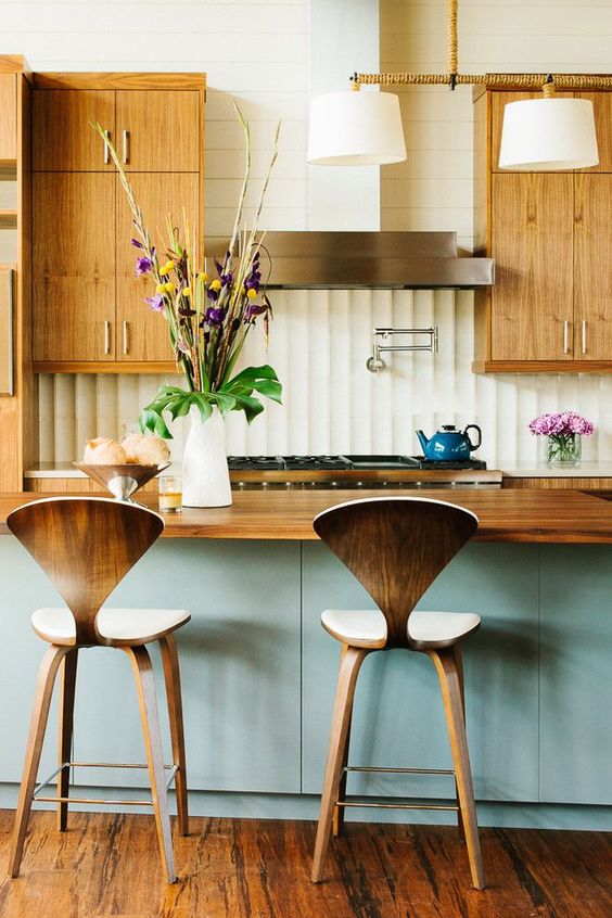 a welcoming mid-century modern kitchen with light-colored cabinets with metallic handles, plywood stools, a blue kitchen island and a wooden countertop