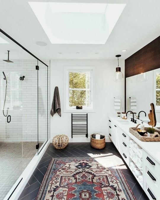 an airy mid-century modern bathroom with white tiles, a skylight, a boho rug, a large vanity with a wooden top and a large shower space with windows