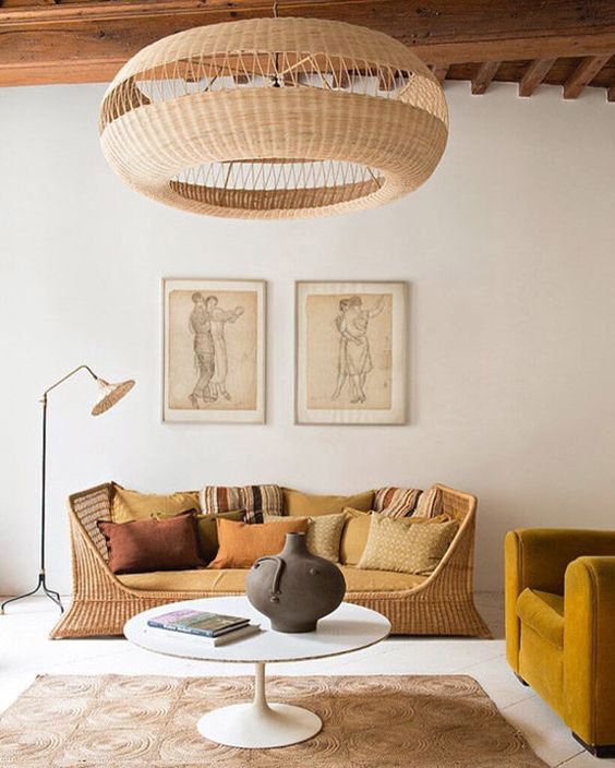 a chic living room with a wicker bench with lots of pillows, a mustard chair, a jute rug and a round table, a wicker pendant lamp