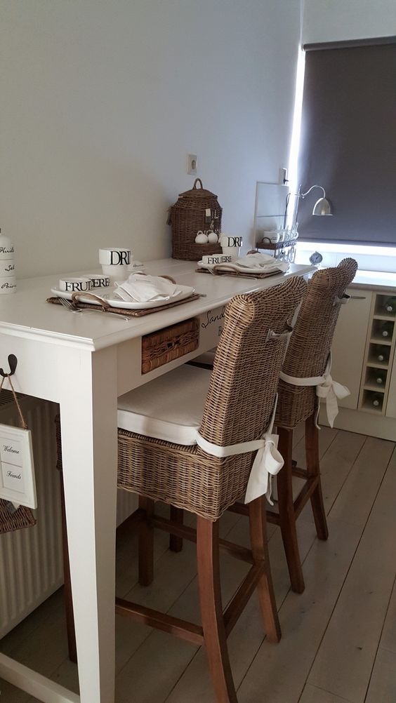 a lovely breakfast nook with a tall white table, tall wicker stools, a wicker basket for storage on the table and lamps around