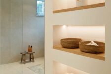a minimalist bathroom with neutral walls, a large shower space enclosed in glass, a series of lit up niche shelves for storage