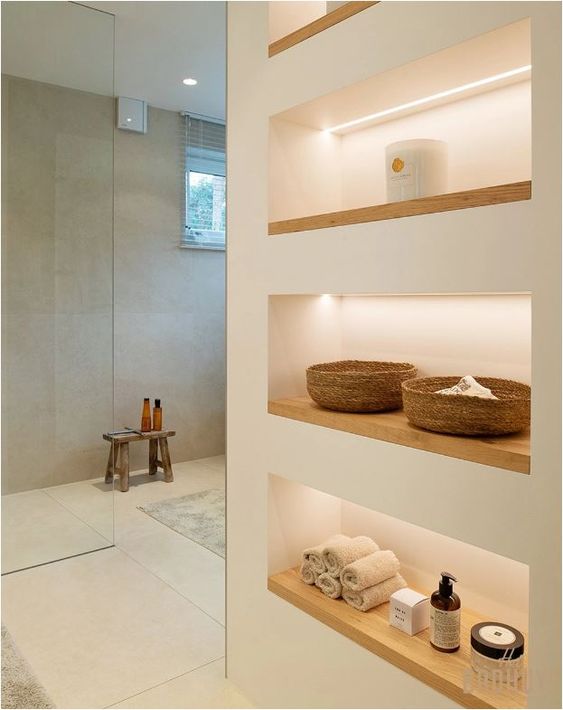 a minimalist bathroom with neutral walls, a large shower space enclosed in glass, a series of lit up niche shelves for storage