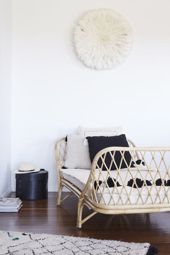 a neutral relaxing nook with a wicker daybed, black and white pillows, a black box for storage and a fluffy lamp on the wall