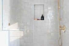 a white shower space with Zellige tiles, a niche shelf, gold fixtures and a small shelf or bench in the corner