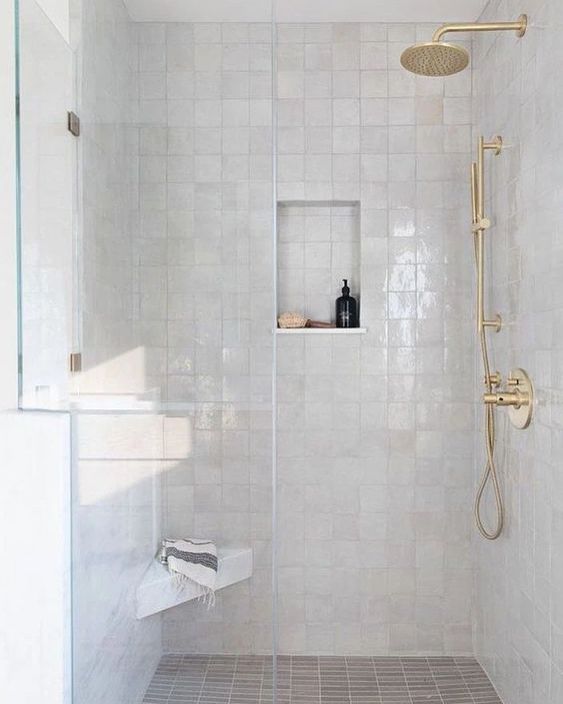 a white shower space with Zellige tiles, a niche shelf, gold fixtures and a small shelf or bench in the corner
