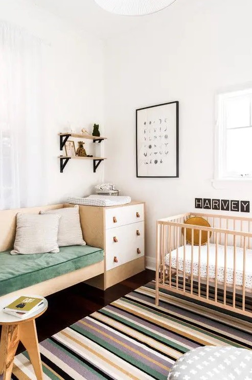 a Nordic chic nursery with white and stained furniture, a built in sofa, a brigth striped rug, open shelves and a printed pouf