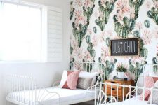 a boho desert shared girls’ bedroom with white metal beds, a jute rug, colored and printed pillows, a cactus print accent wall and a stained dresser