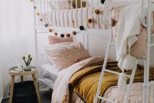 a boho shared girls’ bedroom with a metal bunk bed, pink and mustard bedding, a round side table and pendant lamps