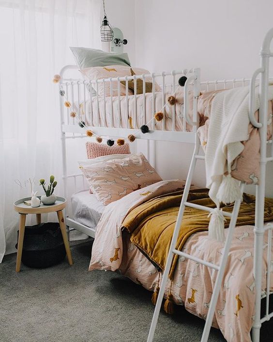 a boho shared girls' bedroom with a metal bunk bed, pink and mustard bedding, a round side table and pendant lamps