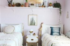 a boho shared girls’ bedroom with light pink walls, white beds and a nightstand, a shelf with lots of plants, a woven pendant lamp