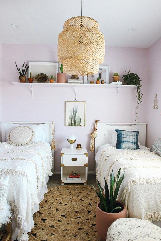 a boho shared girls' bedroom with light pink walls, white beds and a nightstand, a shelf with lots of plants, a woven pendant lamp a boho shared girls' bedroom with light pink walls, white beds and a nightstand, a shelf with lots of plants, a woven pendant lamp