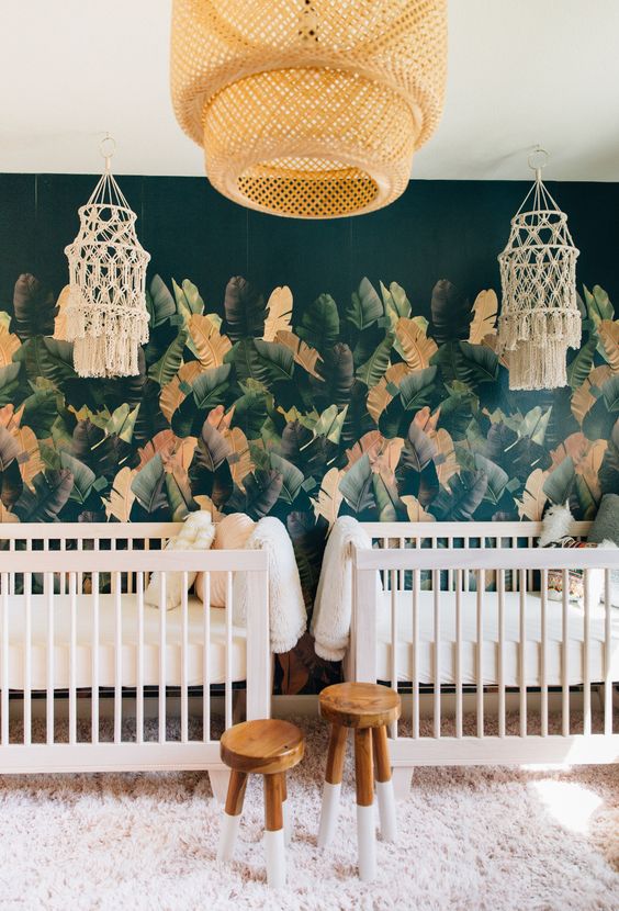 a boho tropical nursery with a banana leaf wall, white and stained furniture, a rattan lamp and crochet lamps over beds