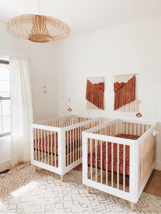 a boho twin nursery in white, with rust and orange touches, with printed bedding, a rattan chandelier and fringe artworks