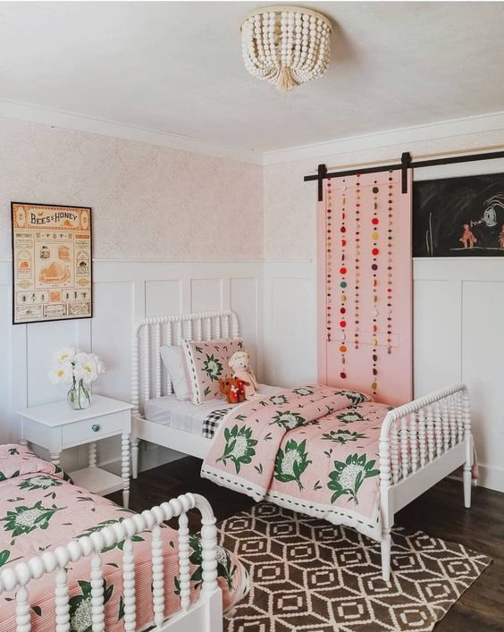a bright shared girls' bedroom with white vintage furniture, pink bedding and a pink barn door with pompoms, a printed rug a bright shared girls' bedroom with white vintage furniture, pink bedding and a pink barn door with pompoms, a printed rug