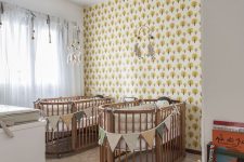 a bright shared nursery with printed wallpaper, stained cribs and neutral furniture, colorful fabric buntings and mobiles