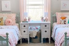 a chic attic kids’ bedroom with mint vintage beds, pastel bedding, a white carved desk and a basket, blush curtains and some pretty decor on the walls