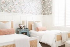 a chic modern shared girls’ bedroom with creamy paneling, white beds with cane, white poufs, neutral and pastel bedding, a floral wallpaper accent