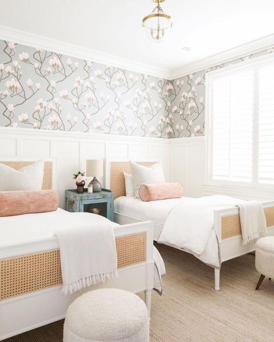 a chic modern shared girls' bedroom with creamy paneling, white beds with cane, white poufs, neutral and pastel bedding, a floral wallpaper accent