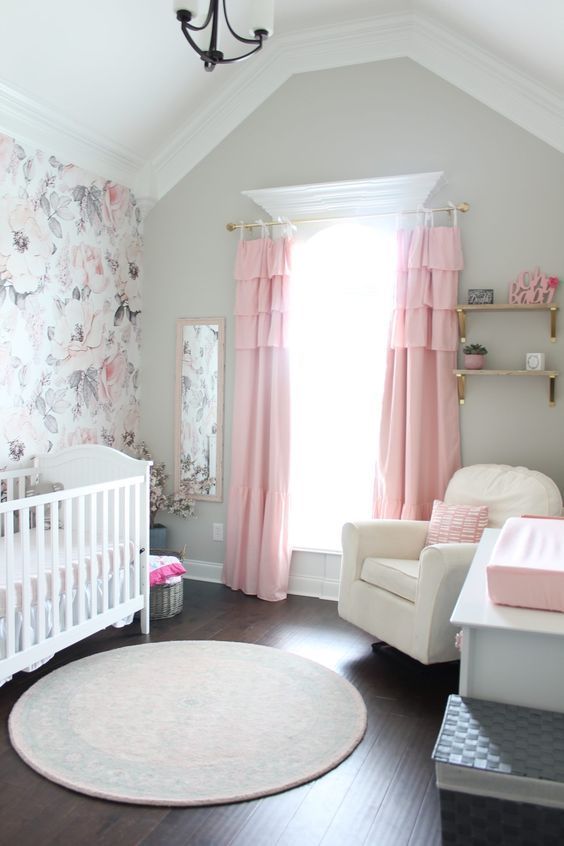 a chic nursery with grey walls, a floral accent wall, white furniture, open shelves, pink tiered curtains, a round rug and pink bedding