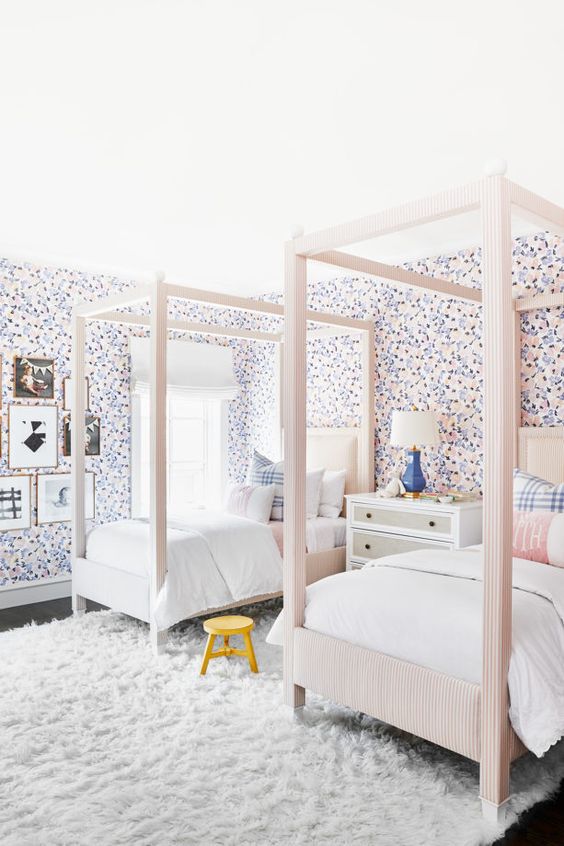 a chic shared girls' bedroom with pastel floral wallpaper, blush upholstered beds, neutral and pastel bedding, a white faux fur rug and a dresser with a lamp, a gallery wall