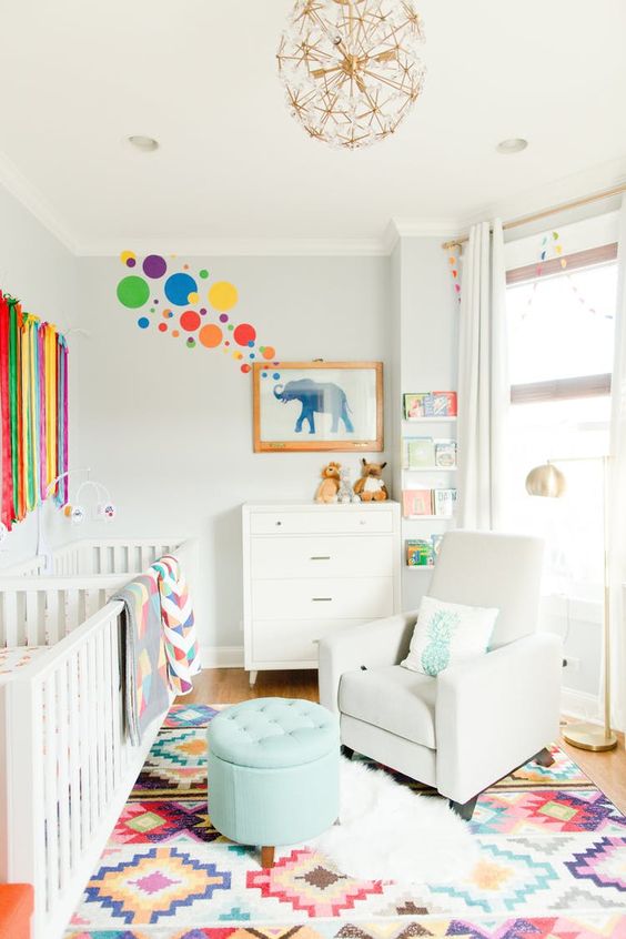 a colorful nursery with a bold wall, a colorful rug and textiles plus a wall hanging and white furniture as a backdrop