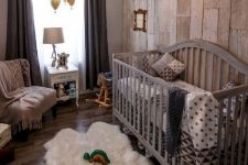 a cool rustic nursery with a reclaimed wood wall, a large grey crib, a fluffy rug, dark curtains and a vintage chandelier