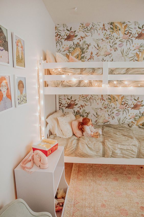 a cozy shared girls' bedroom with a floral accent wall, a bunk bed accent with lights, a white toy chest and a gallery wall a cozy shared girls' bedroom with a floral accent wall, a bunk bed accent with lights, a white toy chest and a gallery wall