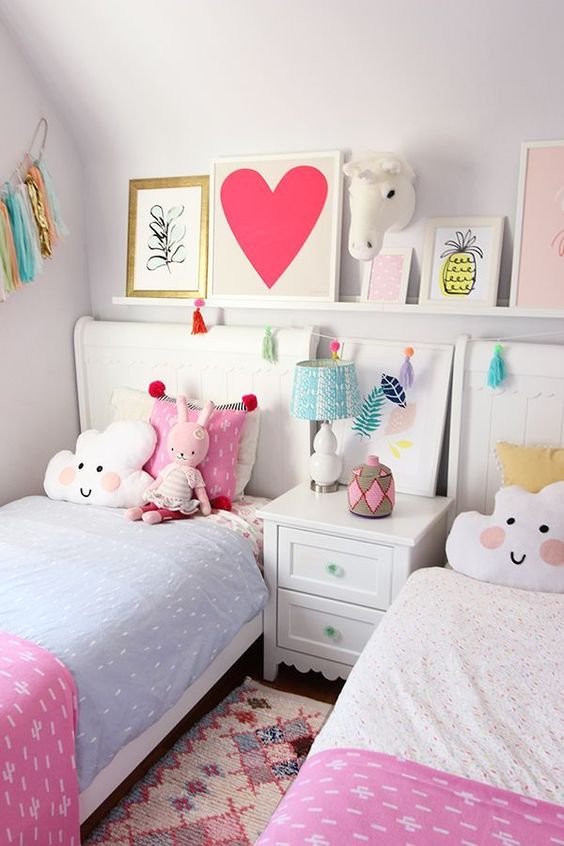 a cute and bright shared girls' bedroom with white furniture, pastel bedding, a ledge gallery wall, a banner and colorful toys and pillows