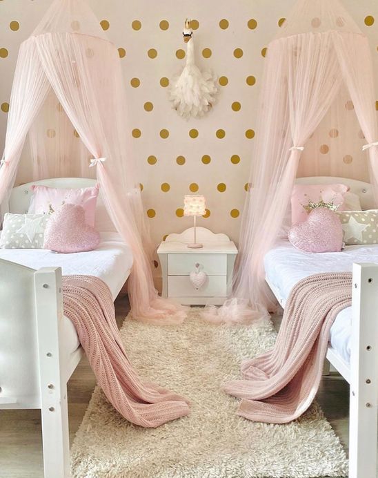 a cute and glam shared girls' bedroom with a gold polka dot accent wall, white beds with white, pink and gold bedding, a gold rug and pink sheer canopies