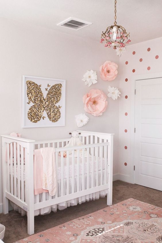 a cute girlish nursery with paper flowers and polka dots on the walls, white furniture, a gold butterfly artwork and a floral chandelier