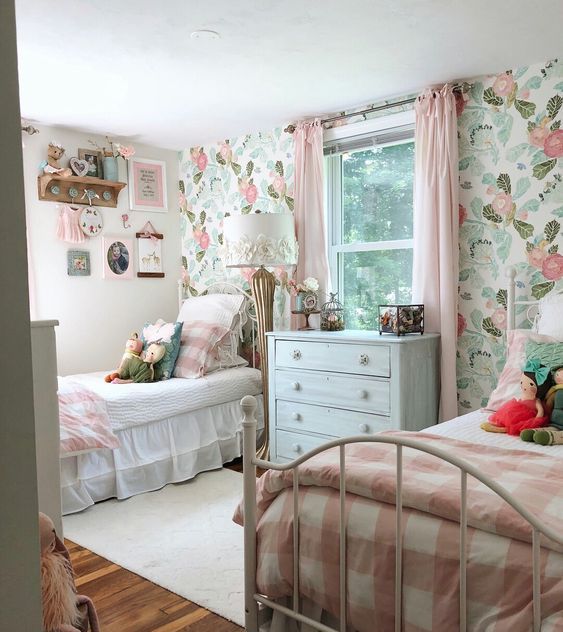 a cute shared girls' bedroom with a pastel floral wall, white metal beds with pink and white bedding, wall-mounted shelves and toys is very cute
