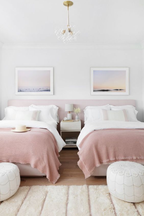 a dreamy shared girls' bedroom with a pink headboard and beds with white and pink bedding, white leather poufs and a nightstand, ocean prints