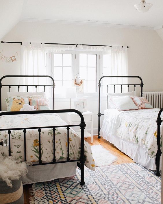 a light-filled shared girls' bedroom with black metal beds with floral bedding, white curtains and a nightstand