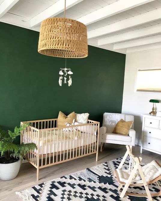 a mid-century modern boho nursery with a green accent wall, white and light-colored furniture, a printed rug and a woven lamp