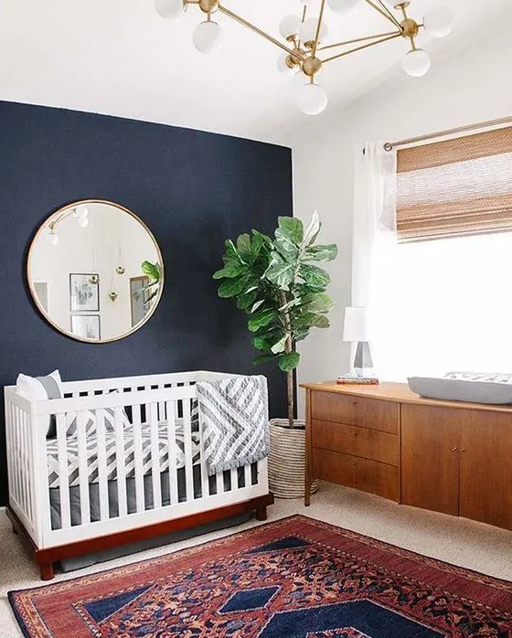 a mid century modern nursery with navy walls, a white crib, a stained dresser, a round mirror, a bright printed rug and a gold chandelier