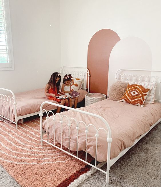 a modern boho shared girls' bedroom with white metal beds, pastel bedding, cool decor on the wall, layered rugs and pillows