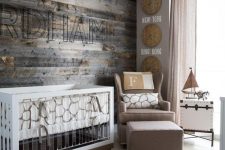 a modern farmhouse nursery with a reclaimed wooden wall, a taupe chair and footrest, a modern crib with acryl and a printed rug