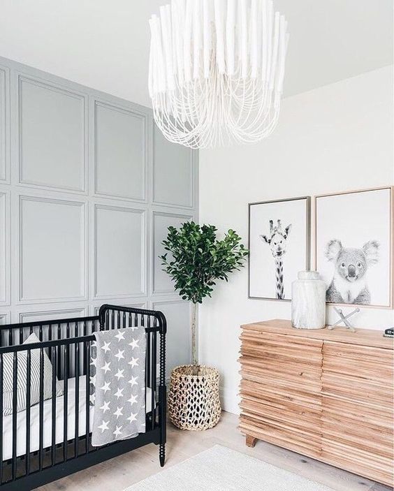 a modern gender neutral nursery with a grey paneled wall, a wooden sideboard, a black crib and a catchy chandelier