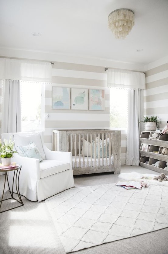 a neutral farmhouse nursery with a whitewashed crib, a unique chandelier, a printed rug and stripes on the walls