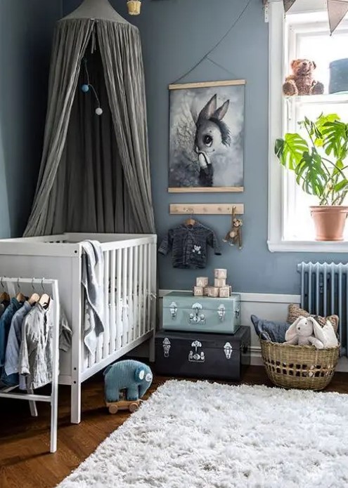 a pastel nursery in Scandi style with blue walls, a white crib, a dreamy artwork, some suitcases and a basket for storage and cool toys