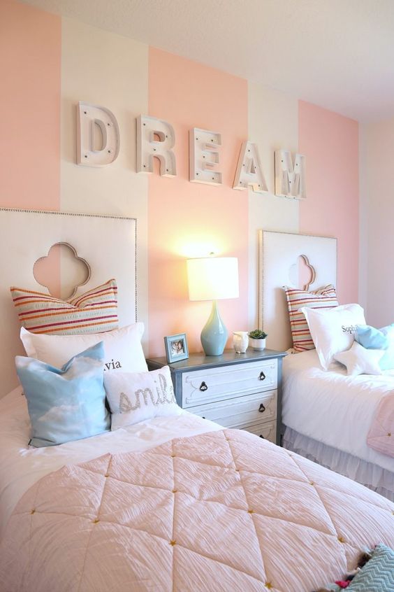 a pastel pink and white girls' bedroom with a striped accent wall, marquee letters, white upholstered beds with pastel bedding, a blue and white dresser a pastel pink and white girls' bedroom with a striped accent wall, marquee letters, white upholstered beds with pastel bedding, a blue and white dresser