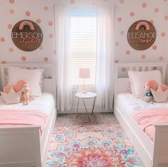 a pretty and bright shared girls' bedroom with a polka dot accent wall, white farmhouse beds, pink and white bedding, a colorful rug and names