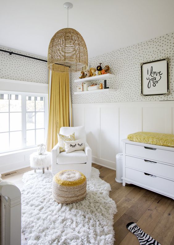 a pretty gender neutral nursery with a wallpaper and panel wall, white furniture, a woven lamp and touches of bright yellow