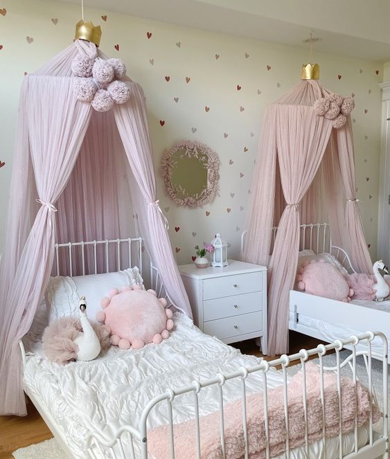 a princess-style shared girls' bedroom with a white metal bed, pink canopies, pink pillows, a white nightstand and a mirror in a pink frame